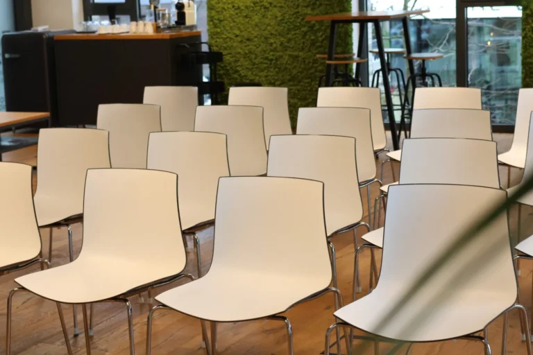 Weiße Stühle in Reihenbestuhlung im Seminarraum mit Blick auf die Kaffee-Ecke und Pausenbereich
